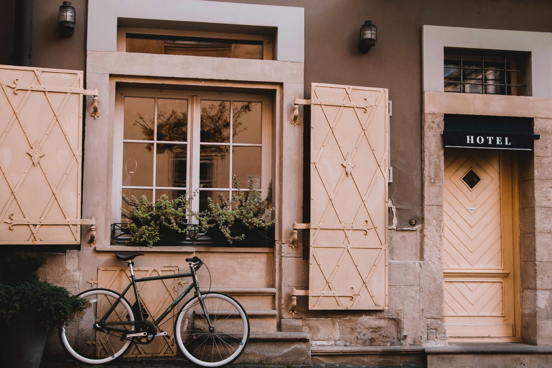 The hotel's shuttered facade at dusk, with a lantern glowing beside the door and a bicycle leaning under a flower-boxed window.