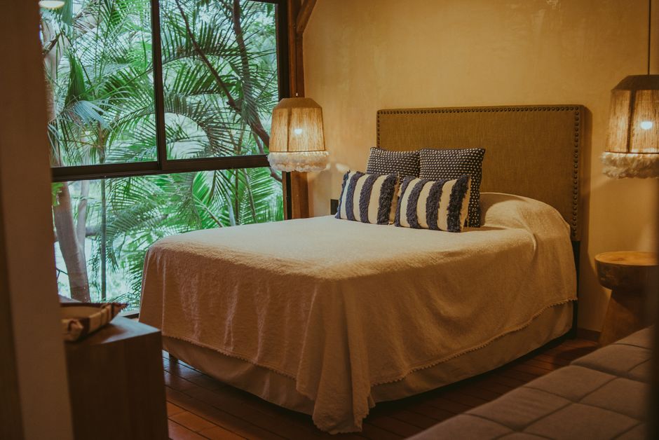 A snug queen bedroom with warm timber floors, rattan pendant lamps and tropical greenery framed in the window.